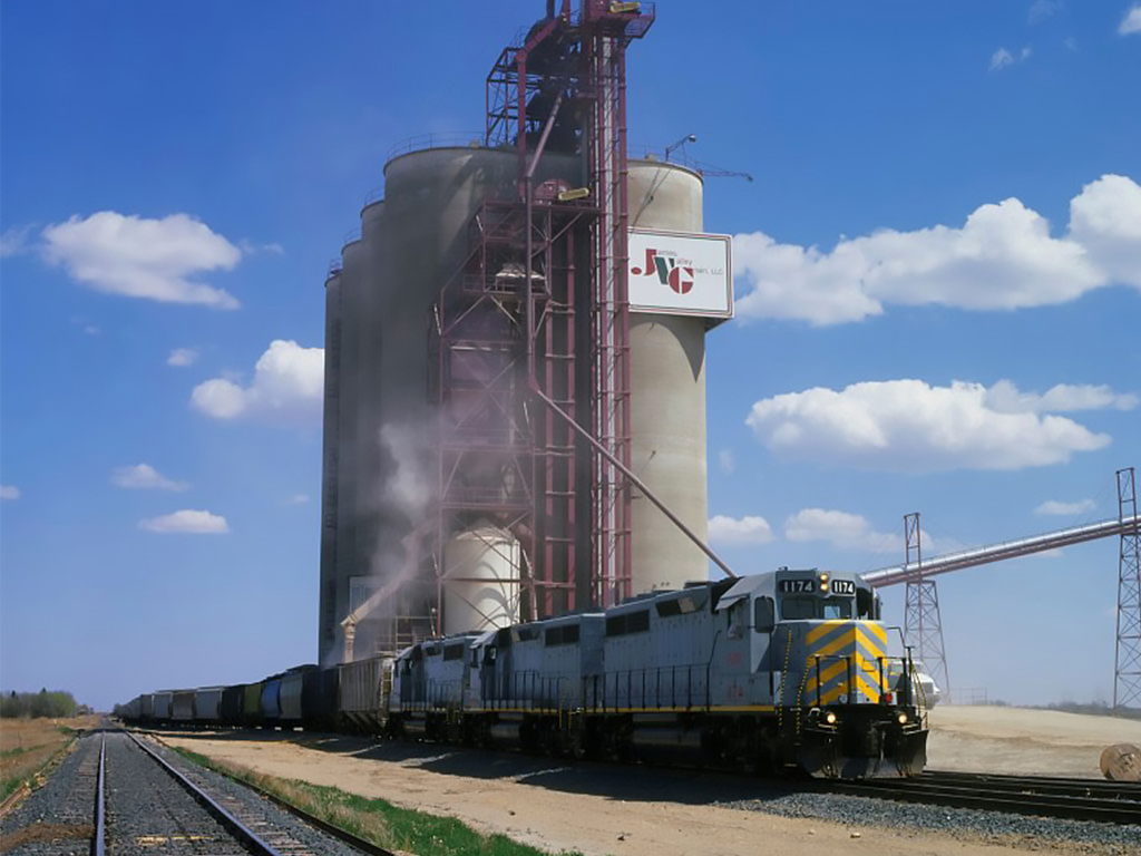 Freight train locomotive pulling cargo cars near large agricultural silos with a bucket elevator system under blue sky.
