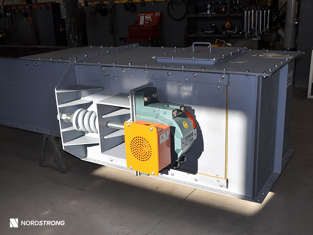 Industrial drag conveyor with exposed shaft and motor drive assembly, featuring a green gearbox and orange safety cover, in a workshop setting.