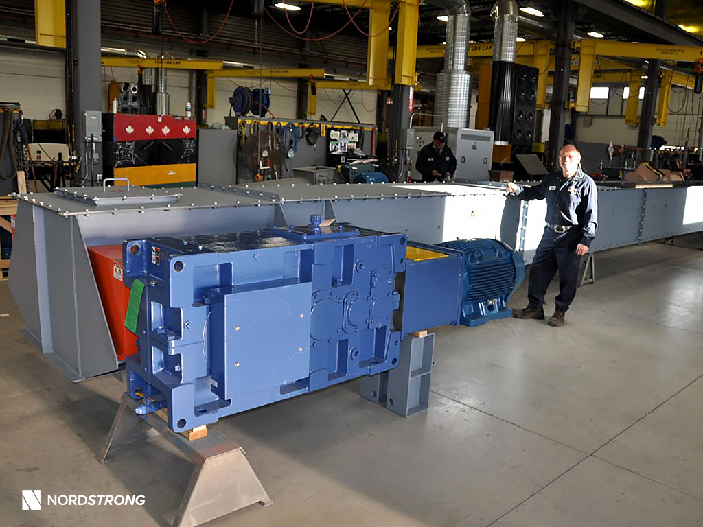 Industrial drag conveyor with large blue gearmotor, shown in a workshop with a technician standing beside the assembled unit.