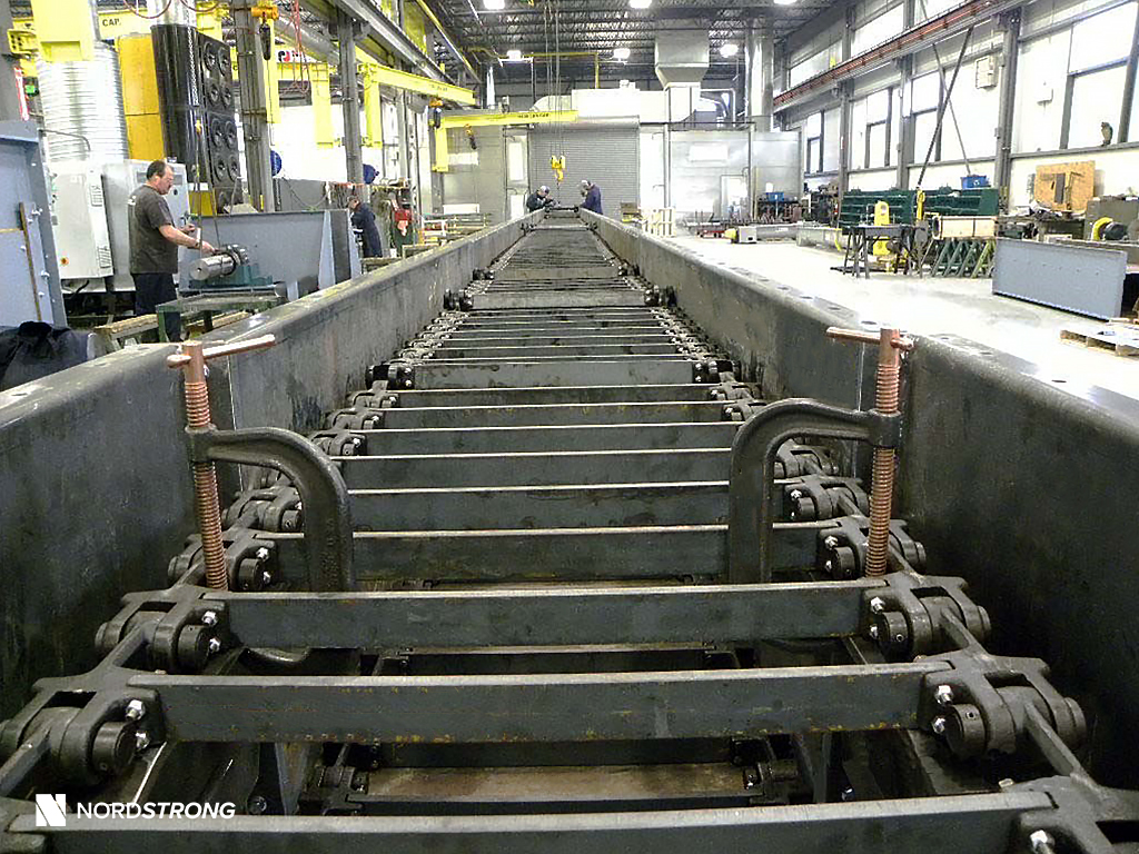Industrial drag conveyor with large blue gearmotor, shown in a workshop with a technician standing beside the assembled unit.