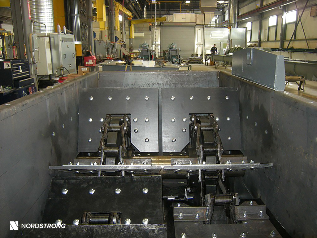 Interior view of an industrial drag conveyor showing heavy-duty chain assembly and paddles, inside a fabrication shop.